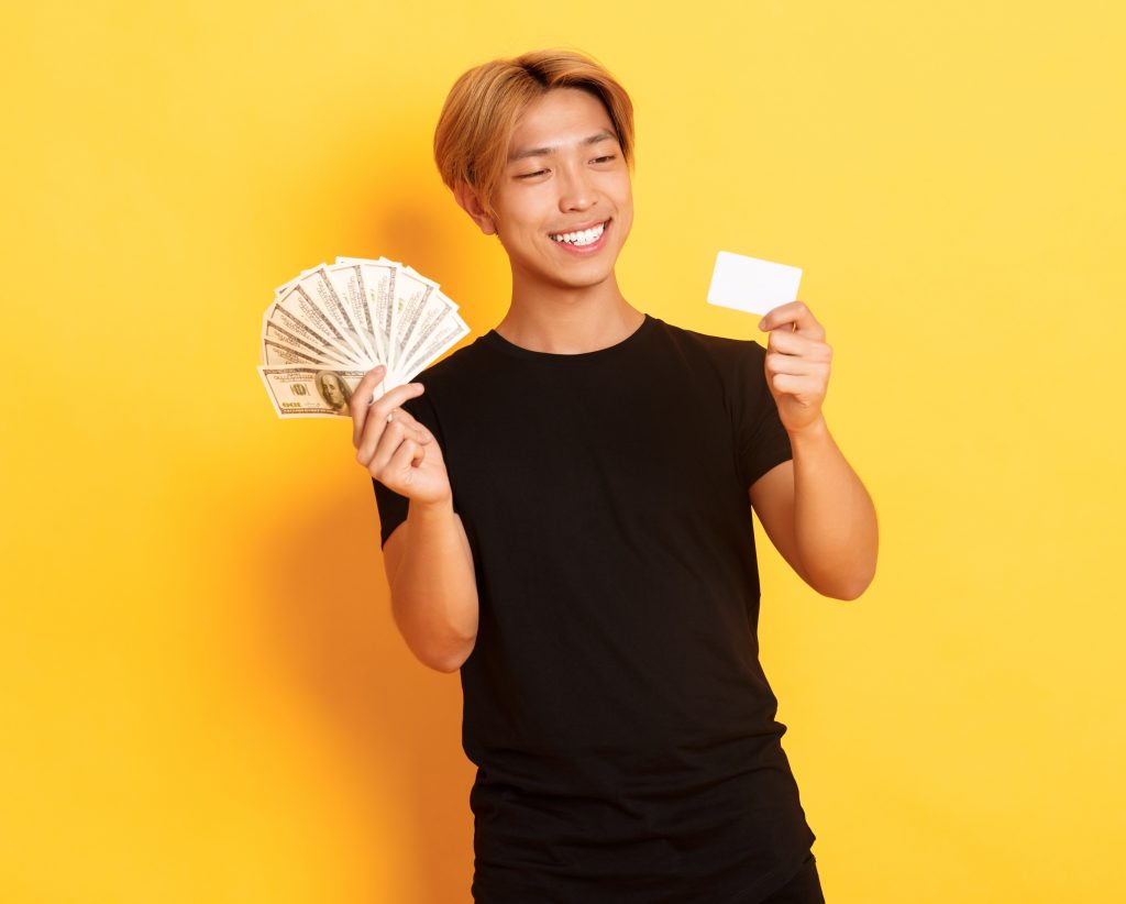asian guy looking at credit card while also holding a money fan, yellow background.